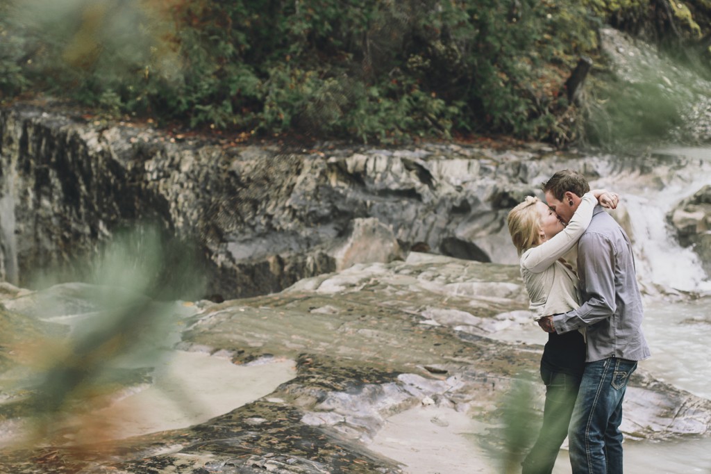 newly engaged white couple snuggles up and kisses against the backdrop of churning river in this engagement session in golden with lolo and noa