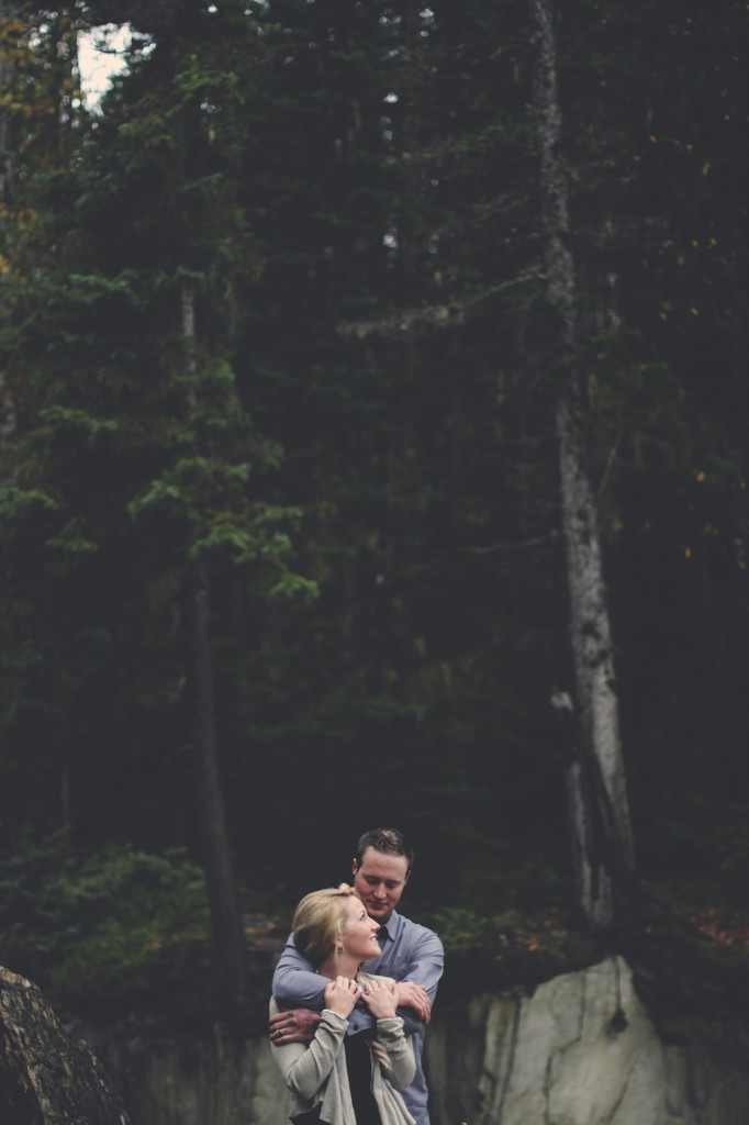 engaged man hugs her fiance's shoulders as she looks back at him smiling against a forest backdrop