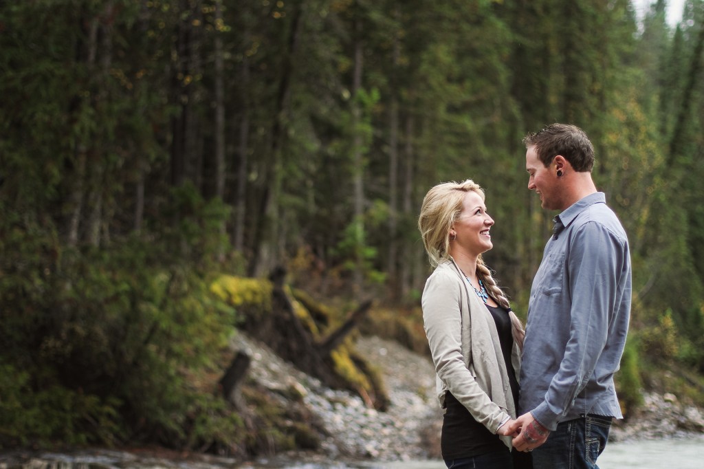 blonde woman with long braid holds her fiance's hand and smiles against a forested backdrop in this golden bc engagement session