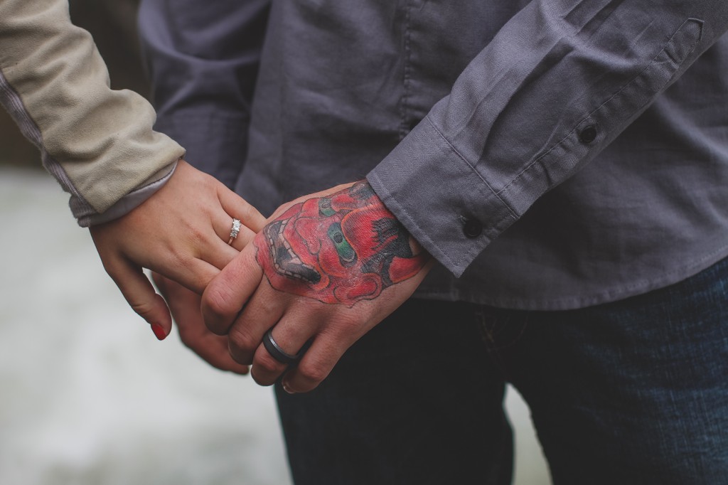 newly engaged couple holds hands to show off their rings and the back of his hand has a red and green tattoo covering it