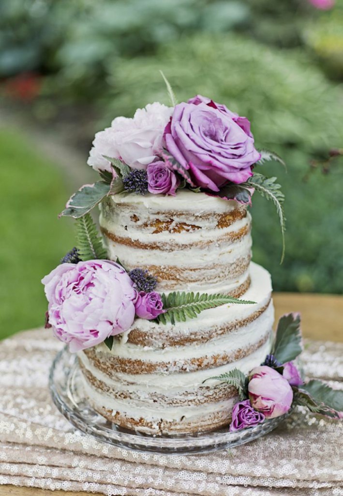 a naked cake sits outside on a table runner with purple roses on top to decorate it