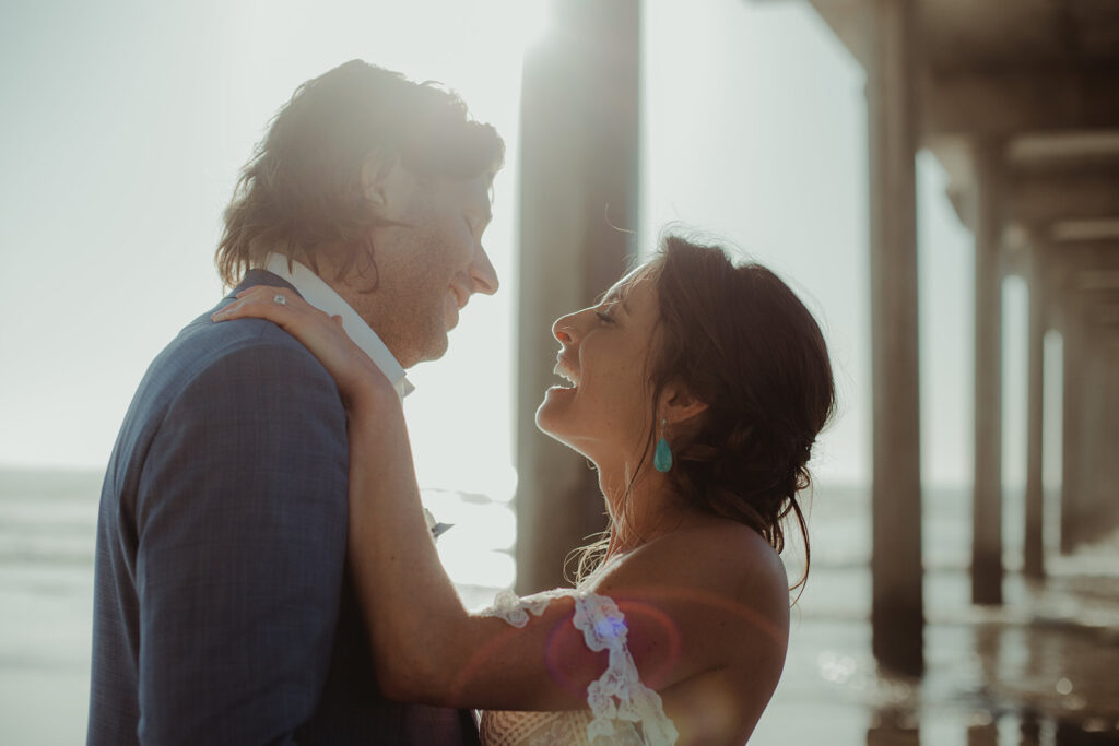 newly wed bride wearing off shoulder casual gown holds onto her groom under a pier on the beach and smiles during this bohemian inspired wedding