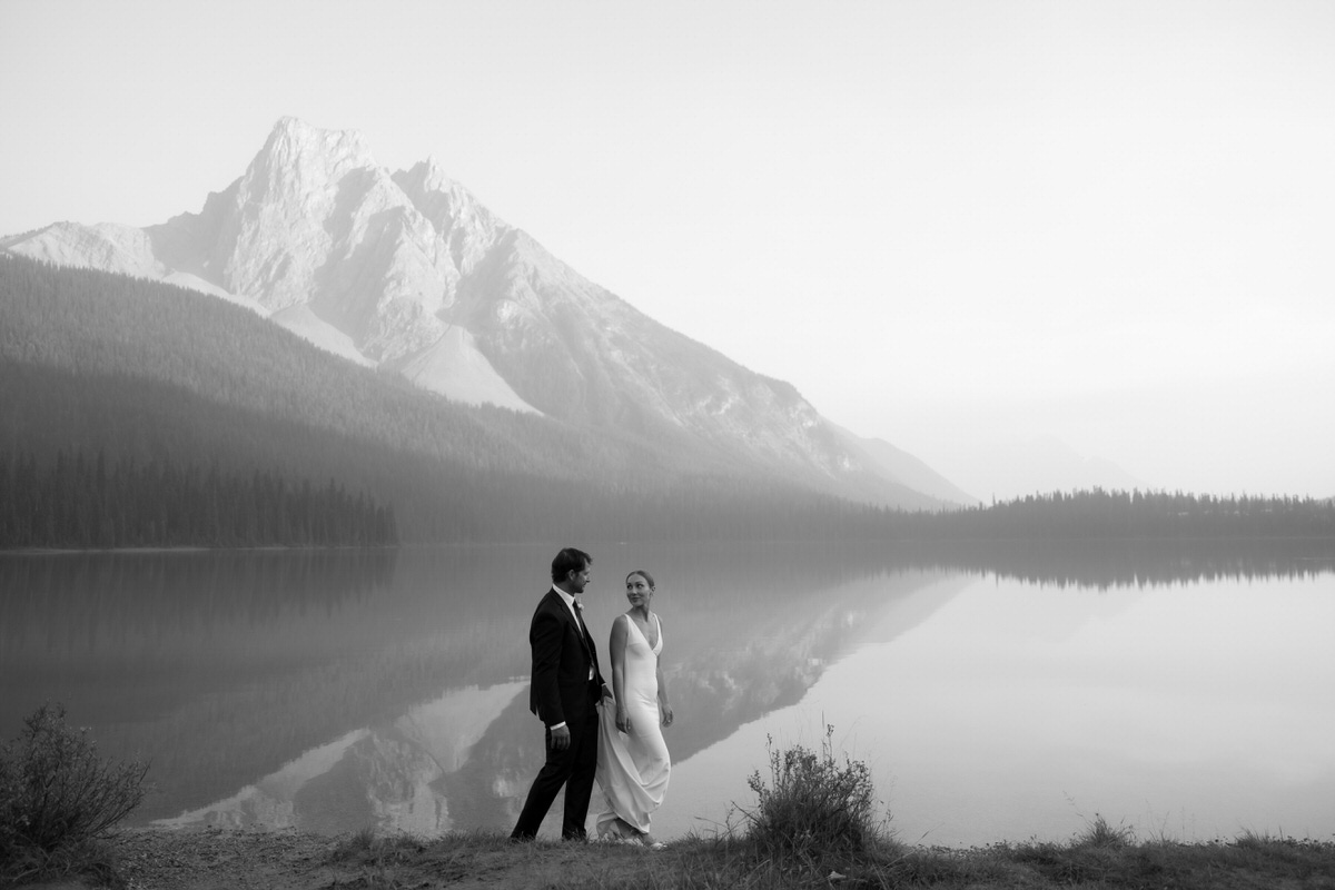 groom carrying bride's train and the bride is looking back at him while they walk along the lake and mountains